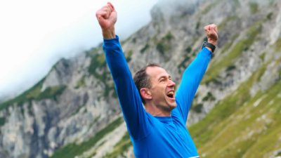 An excited man stretches his arms out above his head as he reaches a mountain peak.