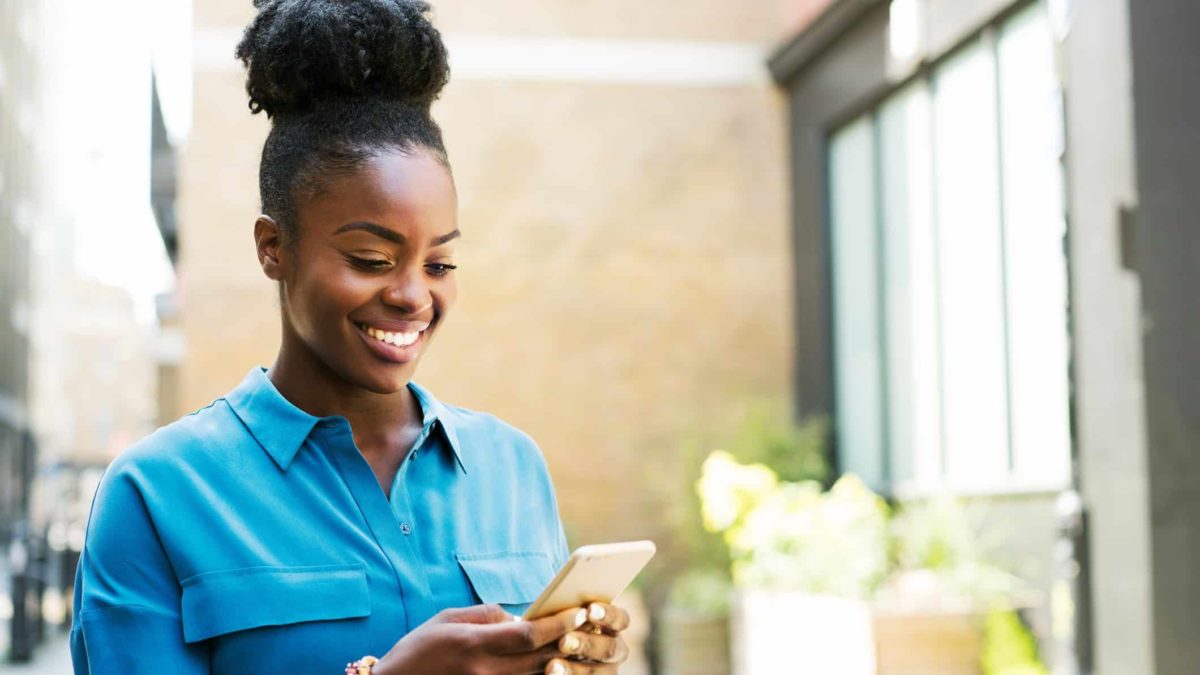 A woman standing in a blue shirt smiles as she uses her mobile phone.
