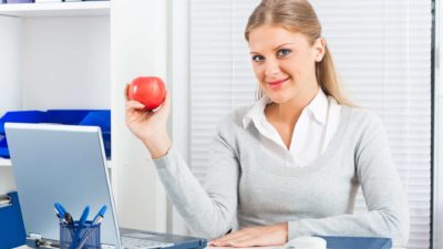 A smiling woman holds up an apple with a laptop open on her desk.