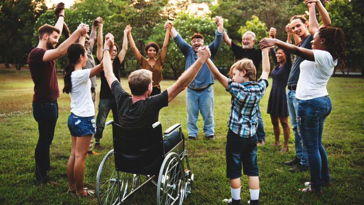 A diverse group of people form a circle at a park and raise their arms together.